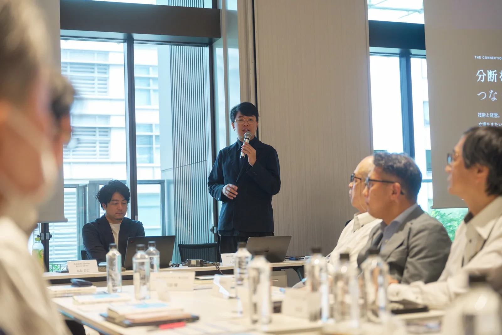 企業の役員が囲むテーブルの前に立ち、マイクを持って話す小澤健祐。スクリーンに「分断を超え、つなぐ」